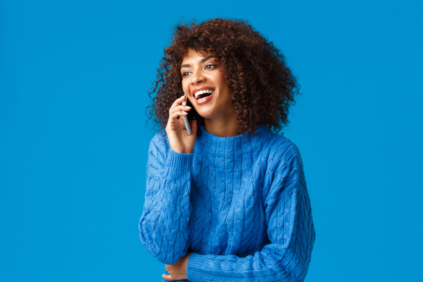 Charismatisch aantrekkelijke gelukkig Afrikaans-Amerikaanse vrouw die de telefoon opneemt, vrolijk en glimlachend lacht, smartphone dicht bij het oor houdt, blauwe achtergrond vrolijk staat, gelukkige feestdagen wenst - Foto, afbeelding