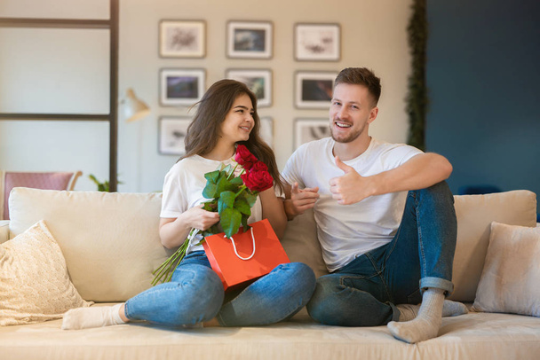 lovely couple hugging on the sofa, handsome husband shows like surprising his beautiful wife with roses and gift on saint valentine's day, happy romantic anniversary - Photo, Image