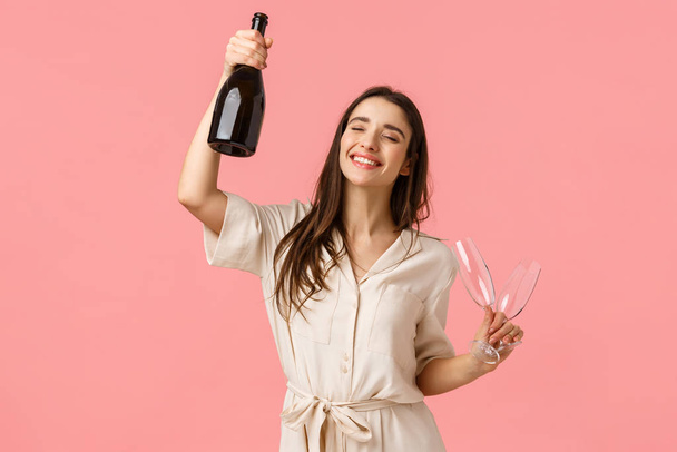 Party, holidays and celebration concept. Cheerful lucky young brunette girl having fun, dancing raising bottle champagne and glass, close eyes and smiling carefree, standing pink background - 写真・画像