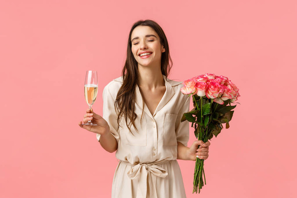 Feier, Party und Schönheitskonzept. Romantisch schöne junge Frau im Kleid, Blumen in der Hand, genießt das Feiern, trinkt Champagner aus Glas, schließt die Augen und lacht, rosa Hintergrund - Foto, Bild