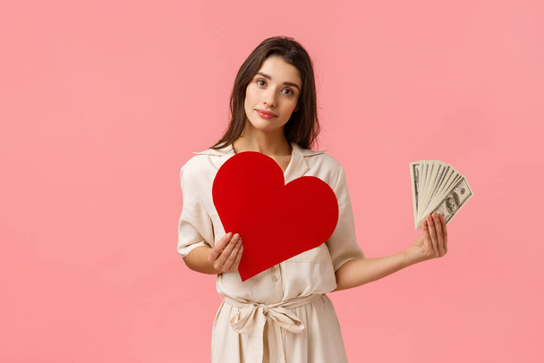 Decisions, relationship and life concept. Alluring romantic young woman having decision made, holding heart card near chest and showing money, tilt head smiling, making choice, pink background - Fotoğraf, Görsel