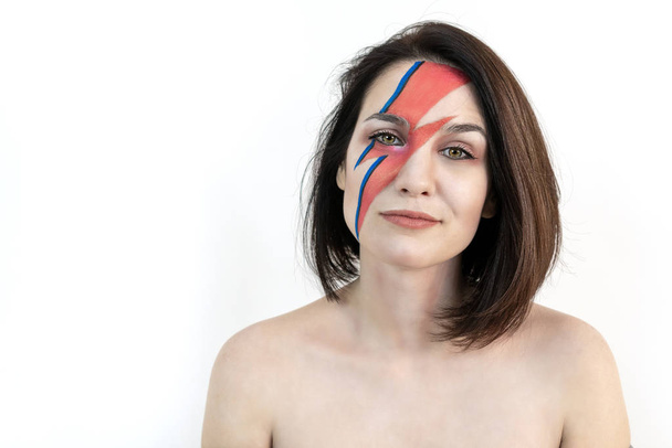 red and blue lightning bolt on her beautiful face. Beautiful girl with original make-up on a white background. Studio photo - Foto, afbeelding