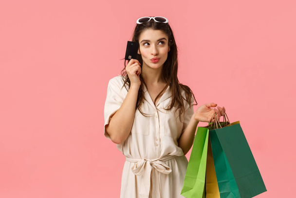Cute glamour, brunette female in dress carrying shopping bags, touching face with credit card, smirk and gazing away thoughtful, imaging what buy next, thinking what else she need, pink background - Fotografie, Obrázek