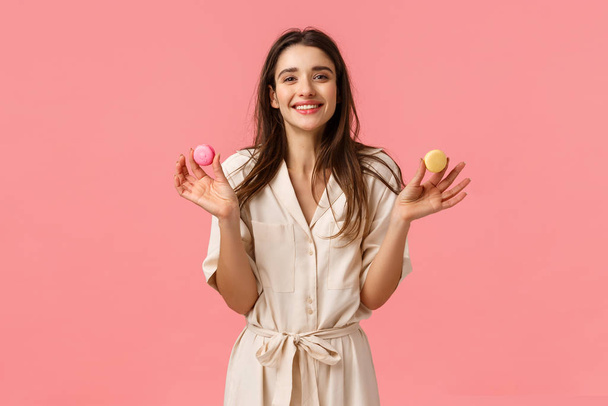 Sweets, happiness and women concept. Cheerful and cute european girl smiling happily, holding two macarons and grinning delighted, sharing dessert with friend, standing pink background - Foto, Imagem