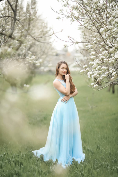 A young stylish model girl in a beautiful fashionable long green dress stands in a summer park near lush flower bushes and looks back - Foto, immagini
