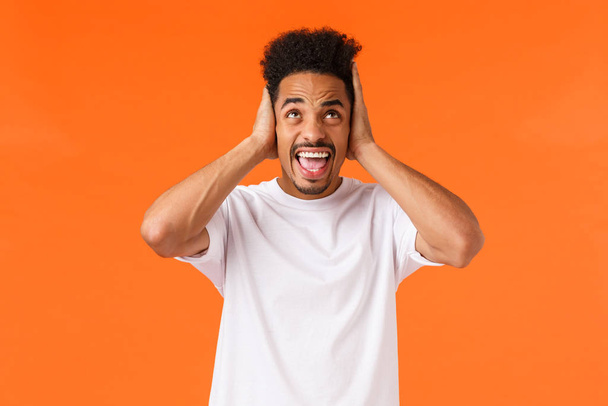 Annoyed and irritated african-american male arguing, complaining noisy neighbours in dormitory, cant study, cover ears, shouting look up displeased and angry, standing orange background - 写真・画像