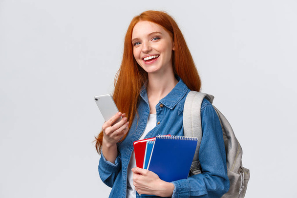 Charismatic, happy smiling redhead teenage girl, college student with backpack, notebooks, holding smartphone, texting friend, chatting or order taxi, waiting for lift grinning camera - Foto, immagini