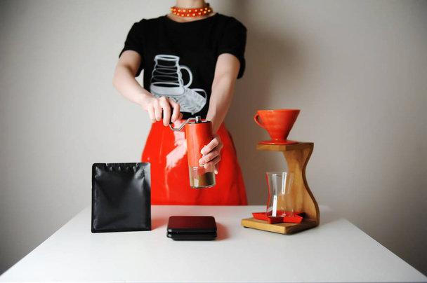 Chica barista de moda elaborando café de filtro. Estación de goteo de madera. Falda de charol rojo, camiseta negra. Amoladora manual
. - Foto, Imagen