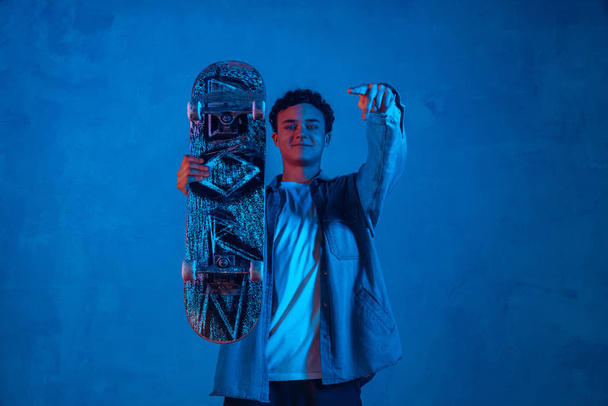Caucasian young skateboarder posing on dark neon lighted background - Фото, изображение