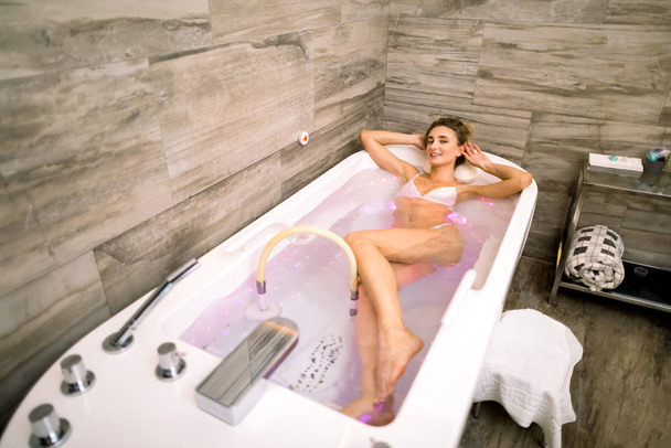 Top angle view of beautiful woman relaxing in the bathtub having a hydromassage therapy in modern spa and cosmetology clinic - Valokuva, kuva