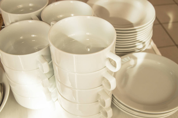 Λευκό κεραμικά καθαρά πιάτα σε στοίβες, που προετοιμάζονται για πρωινό στο ξενοδοχείο, περιμένει τους επισκέπτες, Ηλιόλουστο πρωινό του καλοκαιριού, το φως του ήλιου σε μπολ ζωμό και πιάτα - Φωτογραφία, εικόνα