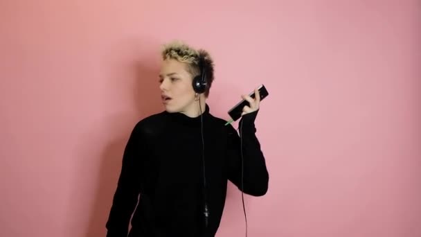 Happy carefree stylish woman in headphones holding smartphone and dancing over pink background - Záběry, video
