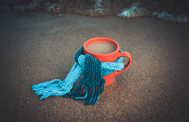 Rode mok met koffie gebonden met blauwe gebreide sjaal staat op zandstrand - Foto, afbeelding