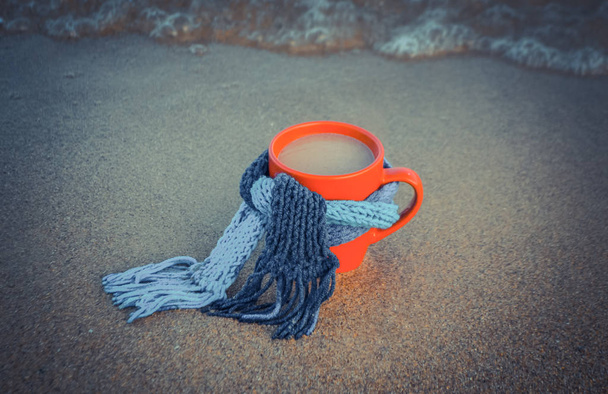 Taza roja con café atado con paños de bufanda de punto azul en la playa de arena
 - Foto, imagen