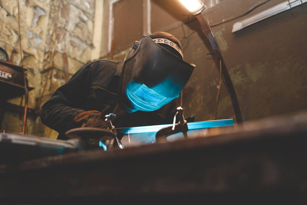 Worker in a welder mask works in a workshop for welding iron. Man makes iron products. Guy works with a welding machine using a mask to protect his eyes from dangerous rays - Foto, imagen