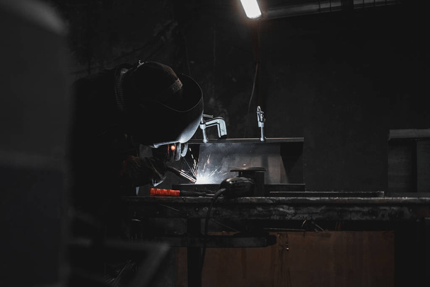 Werknemer in een lasmasker werkt in een werkplaats voor het lassen van ijzer. De mens maakt ijzeren producten. Guy werkt met een lasmachine met behulp van een masker om zijn ogen te beschermen tegen gevaarlijke stralen - Foto, afbeelding