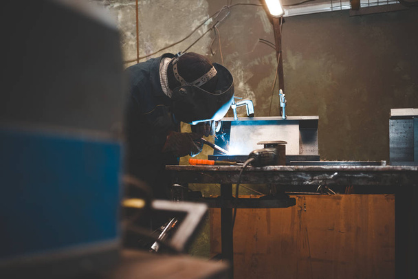 Рабочий в маске сварщика работает в мастерской по сварке утюга. Человек производит железные изделия. Парень работает с сварочной машиной, используя маску, чтобы защитить глаза от опасных лучей
 - Фото, изображение