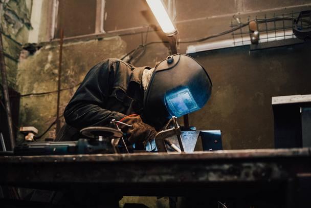 Trabalhador em uma máscara de soldador funciona em uma oficina de soldagem de ferro. O homem faz produtos de ferro. Guy trabalha com uma máquina de solda usando uma máscara para proteger seus olhos de raios perigosos
 - Foto, Imagem