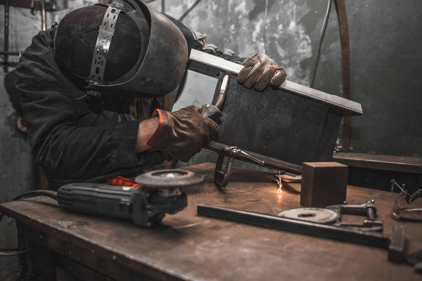 Worker in a welder mask works in a workshop for welding iron. Man makes iron products. Guy works with a welding machine using a mask to protect his eyes from dangerous rays - Photo, Image