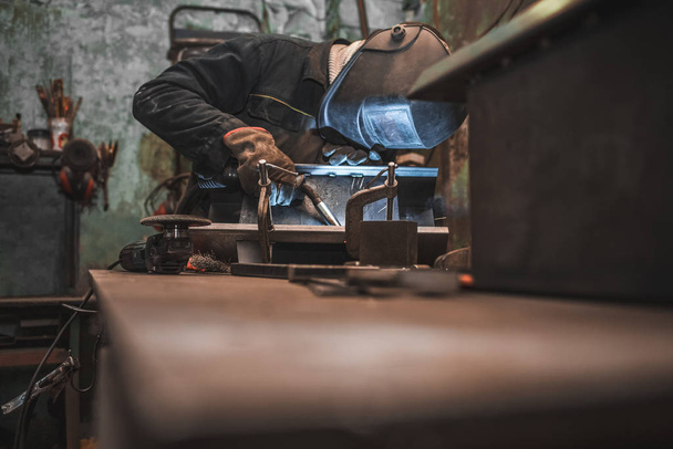 Trabalhador em uma máscara de soldador funciona em uma oficina de soldagem de ferro. O homem faz produtos de ferro. Guy trabalha com uma máquina de solda usando uma máscara para proteger seus olhos de raios perigosos
 - Foto, Imagem