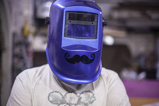Cheerful welder in a welding mask. Mask mustache for eye protection. Master in his workshop. A man at work in the garage. The welder is getting ready for work. A husband in his realm of handicrafts. - Foto, Bild