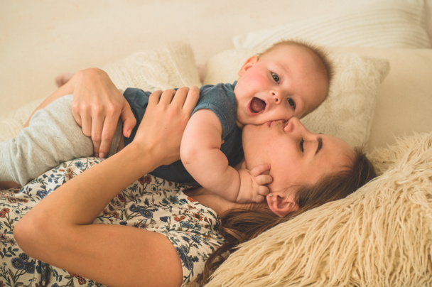 Szeretet és család. Házi portré egy kisfiúról anyával az ágyon. Anya játszik és megcsókolja a gyerekét. - Fotó, kép