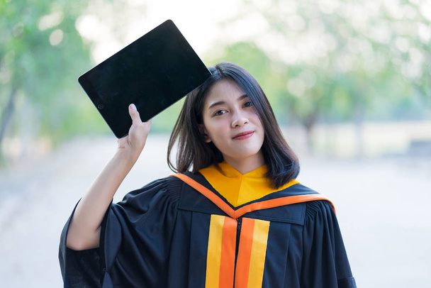 Una joven alegre hermosa graduada con vestido académico y la celebración de la tableta de preparación para la ceremonia de graduación al aire libre
. - Foto, Imagen