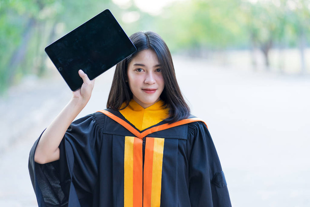 Una joven alegre hermosa graduada con vestido académico y la celebración de la tableta de preparación para la ceremonia de graduación al aire libre
. - Foto, imagen