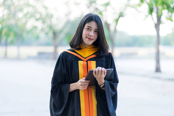 A young cheerful beautiful female gradute wearing academic gown and holding tablet preparing for graduation ceremony outdoor. - Photo, Image
