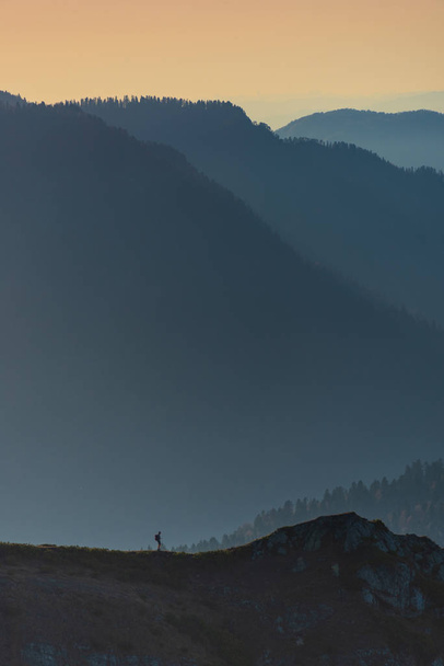 Kleine eenzame wandelaar op de top van een heuvel met een achtergrond van bos bedekte bergen en dalen bij zonsondergang - Foto, afbeelding