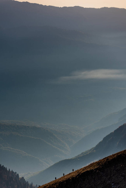 Tre piccoli escursionisti (in basso) che si arrampicano sul fianco di una montagna con una serie di valli lussureggianti, nuvole e cime di montagna alle loro spalle sullo sfondo
  - Foto, immagini