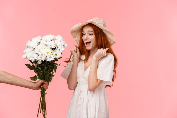 Happy moments, romance and relationship concept. Joyful and surprised cute, lovely redhead girl jumping from happiness and amazement, looking at hand giving her beautiful flowers, pink background - Zdjęcie, obraz
