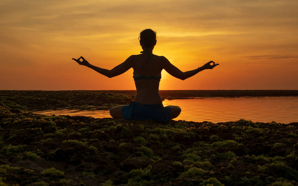 Stanowisko do jogi. Kobieta siedząca na plaży, ćwicząca jogę. Młoda kobieta podnosząca rękę z gyan mudra podczas zachodu słońca złotej godziny. Widok z tyłu. Plaża Melasti, Bali.  - Zdjęcie, obraz