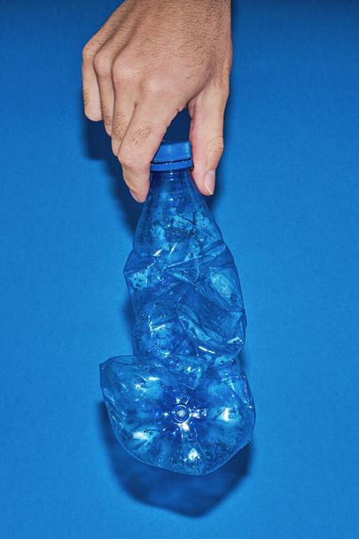 Hand hold squeezed plastic bottle on blue background. Zero waste concept - 写真・画像