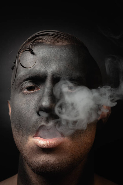 Close-up portrait of a handsome guy in makeup blows smoke. Stock art photo of a smoker. - Zdjęcie, obraz