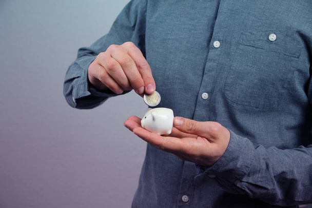 man in a gray shirt puts a 2 euro metal coin into a small white piggy bank, a concept of saving and saving money, banking - Photo, image