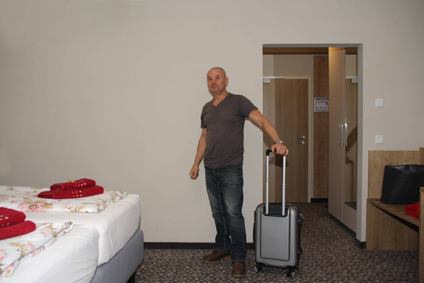 adult man with a suitcase stands in a hotel room and looks at the bed, concept of vacation, business trip - 写真・画像