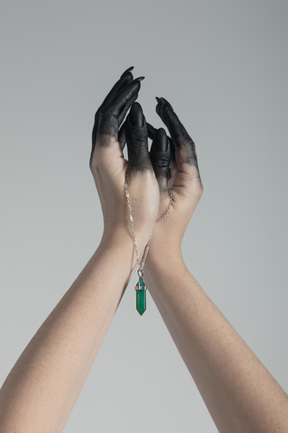 Cropped view of witch with black paint on hands holding green crystal on chain isolated on grey  - Фото, изображение