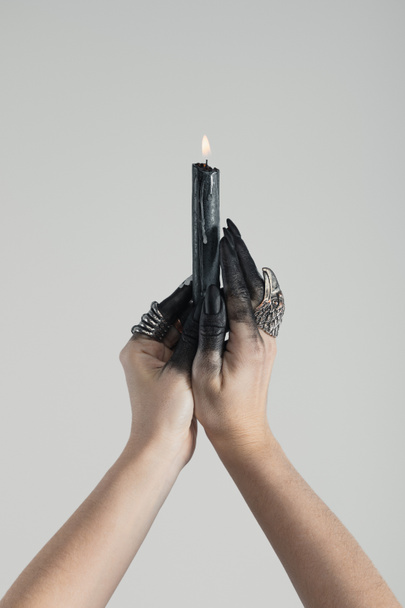 Vista cortada de bruxa com tinta preta nas mãos segurando vela ardente isolada em cinza
 - Foto, Imagem