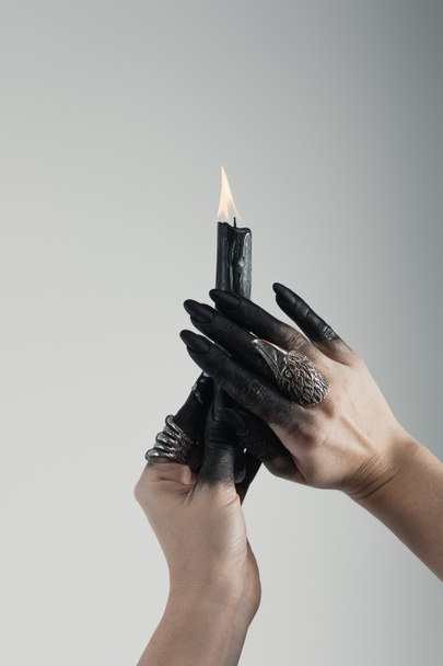 Обрезанный вид рук ведьмы с черными покрашенными пальцами и кольцами ювелирных украшений, держащих свечи изолированы на сером
 - Фото, изображение