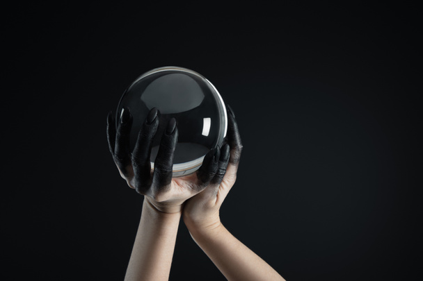 Oříznutý pohled na čarodějnici s černou barvou na ruce držící křišťálovou kouli izolovanou na černé - Fotografie, Obrázek