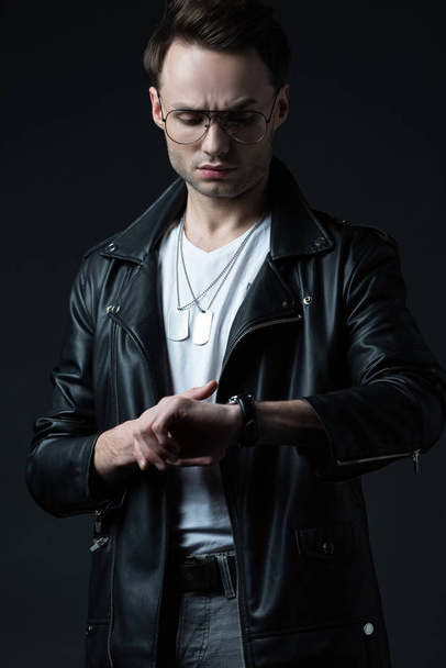 tense stylish brutal man in biker jacket looking at wristwatch isolated on black - Foto, immagini