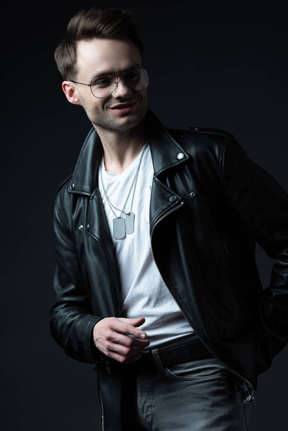 smiling stylish brutal man in biker jacket pointing with finger aside isolated on black - Photo, Image