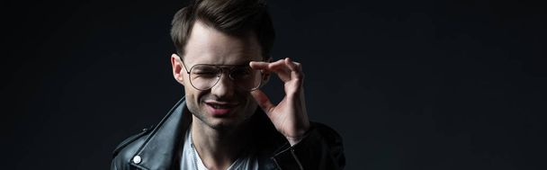 smiling stylish man in biker jacket touching eyeglasses and looking at camera isolated on black, panoramic shot - Fotó, kép