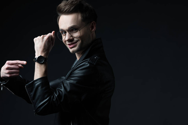 side view of smiling stylish brutal man in biker jacket pointing with finger at wristwatch isolated on black - Photo, Image