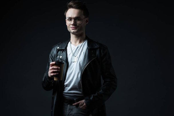 smiling stylish brutal man in biker jacket with hand in pocket holding coffee to go isolated on black - Photo, Image
