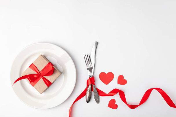 Romantikus asztal beállítás fehér háttérrel. Valentin napi kártya sablon. Kraft ajándékdoboz piros szalaggal a tányéron, ezüst étkészlet. Koncepció évforduló, anyák napja, születésnapi köszöntés, copyspace, felülnézet - Fotó, kép