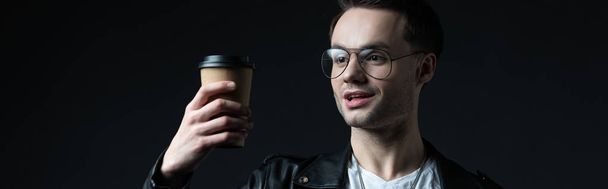 excited stylish brutal man in biker jacket looking at coffee to go isolated on black, panoramic shot - Photo, Image