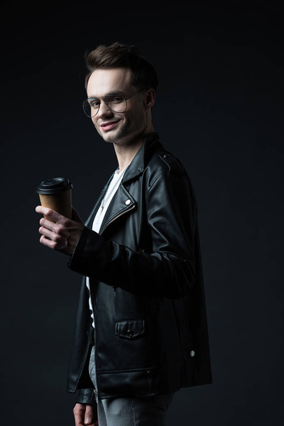 side view of smiling stylish brutal man in biker jacket with coffee to go isolated on black - Fotó, kép