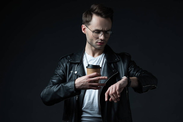 stylish brutal man in biker jacket looking at wristwatch with coffee to go isolated on black - Photo, Image